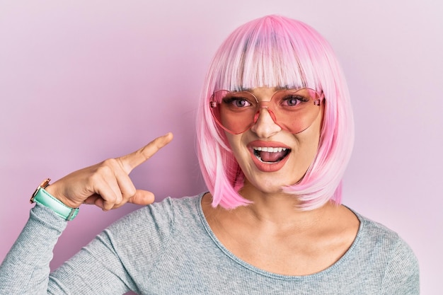 young caucasian woman wearing pink wig and heart sunglasses smiling happy pointing with hand and finger