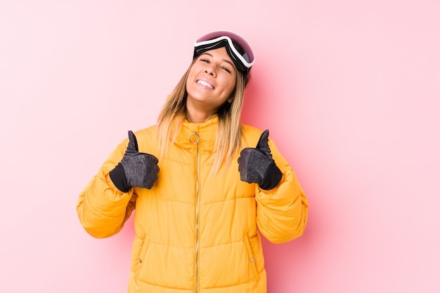 Молодая кавказская женщина нося лыжи одевает в розовой стене поднимая как большие пальцы руки вверх, усмехающся и уверенно.