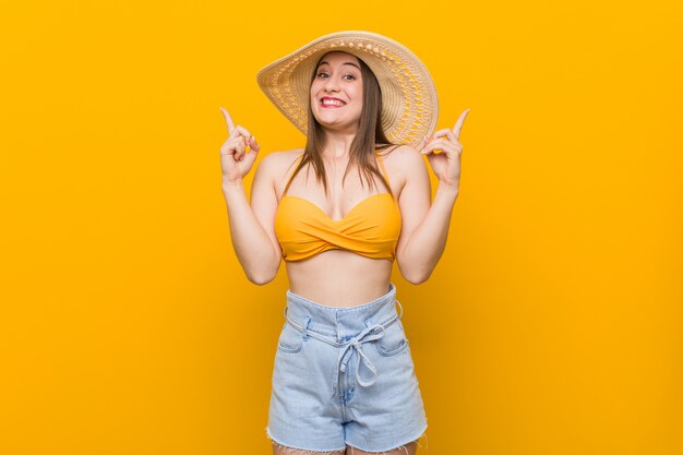 Young caucasian woman wearing a straw hat, summer look indicates with both fore fingers up showing a blank .