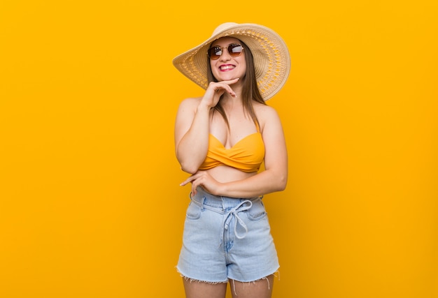 Giovane donna caucasica che indossa un cappello di paglia, look estivo sorridente felice e fiducioso, toccando il mento con la mano.