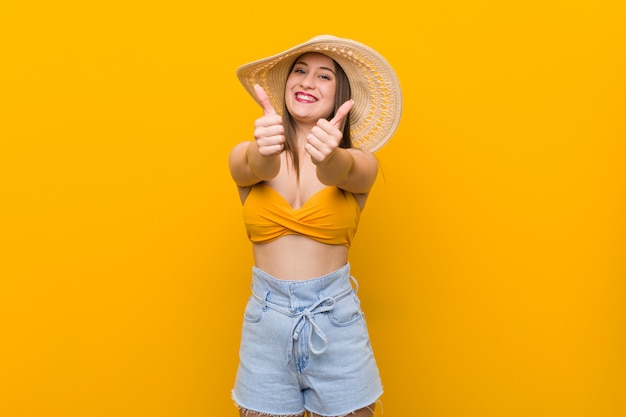 Young caucasian woman wearing a straw hat, summer look with thumbs ups, cheers about something, support and respect concept