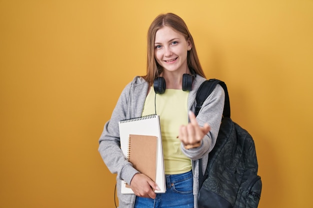 学生のバックパックを着て本を持ち、幸せと笑顔を歓迎する手でここに来てジェスチャーを手招きする若い白人女性