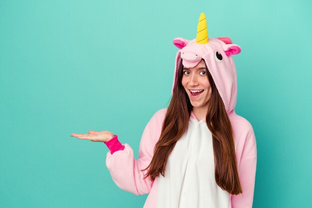 Young caucasian woman wearing a unicorn pajama isolated on blue wall showing a copy space on a palm and holding another hand on waist.