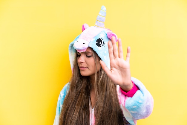 Young caucasian woman wearing a unicorn pajama isolated on\
yellow background making stop gesture and disappointed