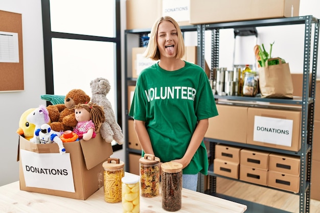 Young caucasian woman wearing volunteer t shirt at donations stand sticking tongue out happy with funny expression. emotion concept.