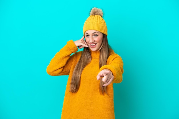 Young caucasian woman wearing winter jacket isolated on blue background making phone gesture and pointing front