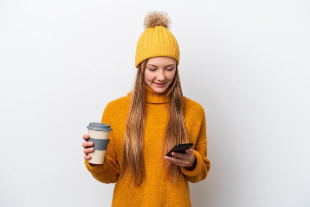 Young caucasian woman wearing winter jacket isolated on white background holding coffee to take away and a mobile