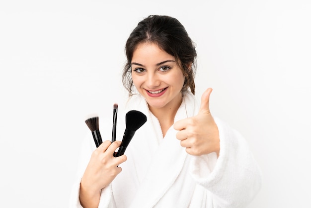 Young caucasian woman on white wall holding makeup brush and with thumb up