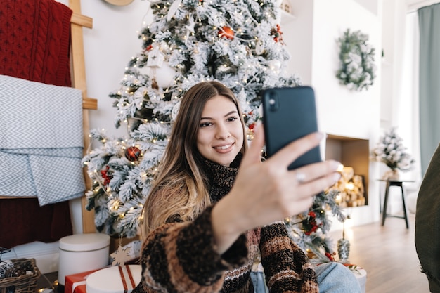 Young caucasian woman wishes merry Christmas by video call using mobile phone in holidays at home on the sofa.