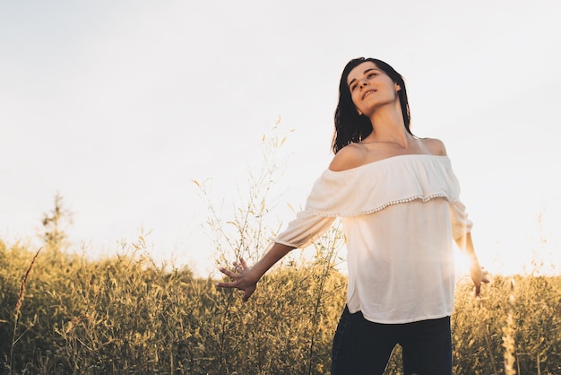 Young Caucasian woman with arms outstretched and palm in field on nature meadow background feeling free Beautiful smiling brunette woman looking at one side on sunset background