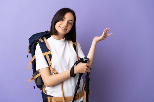 Young caucasian woman with backpack and trekking poles isolated on blue extending hands to the side for inviting to come