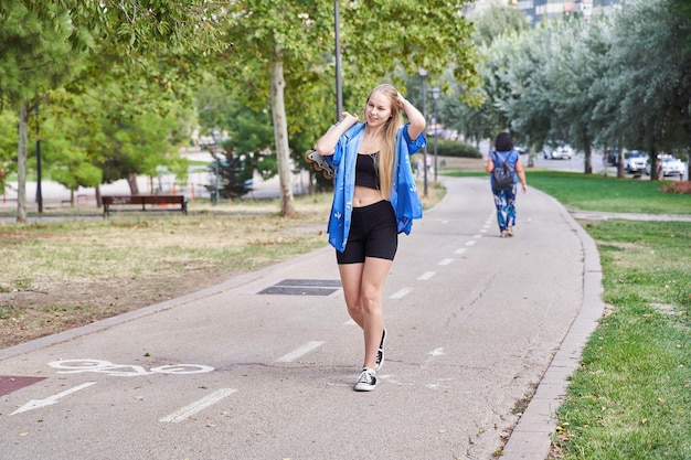 Young caucasian woman with blonde hair walks outdoors in a park with inline roller skates in hand to go to skating training and play sports on a hot sunny day