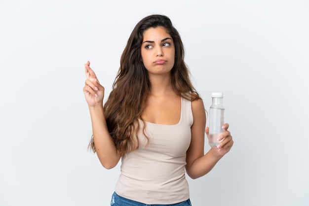 Young caucasian woman with a bottle of water isolated on white background with fingers crossing and wishing the best