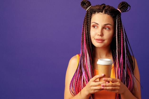 Young caucasian woman with colorful long cornrows holding a cup of coffee against purple background 