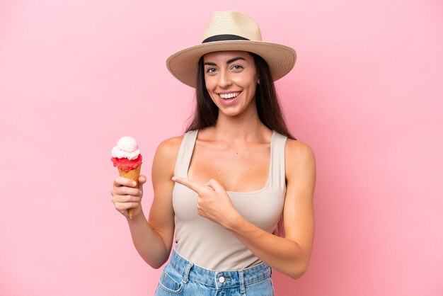 뒤를 가리키는 분홍색 배경에 고립 된 코넷 아이스크림을 가진 젊은 백인 여성