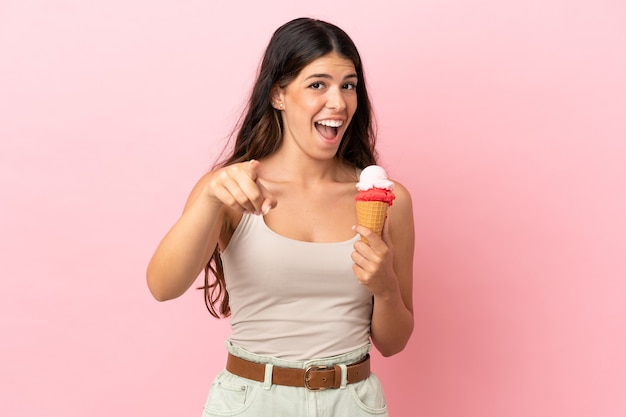 Young caucasian woman with a cornet ice cream isolated on pink background surprised and pointing front