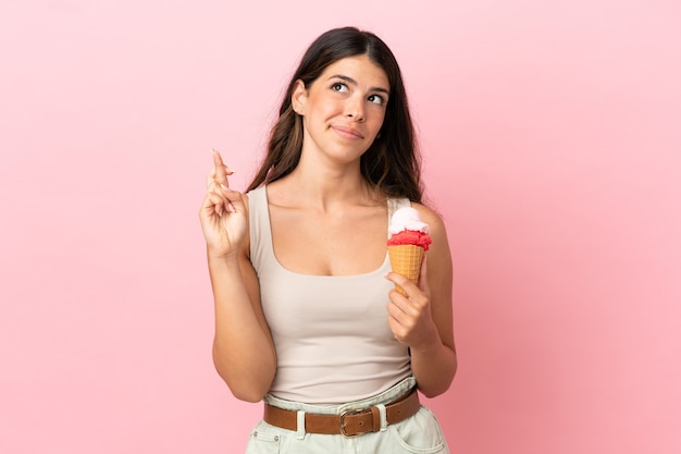 Young caucasian woman with a cornet ice cream isolated on pink background with fingers crossing and wishing the best