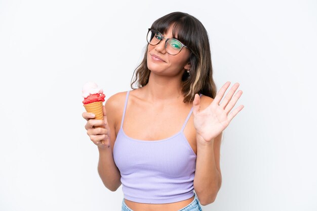 격리된 흰색 배경 위에 코넷 아이스크림을 들고 행복한 표정으로 손으로 경례하는 백인 젊은 여성