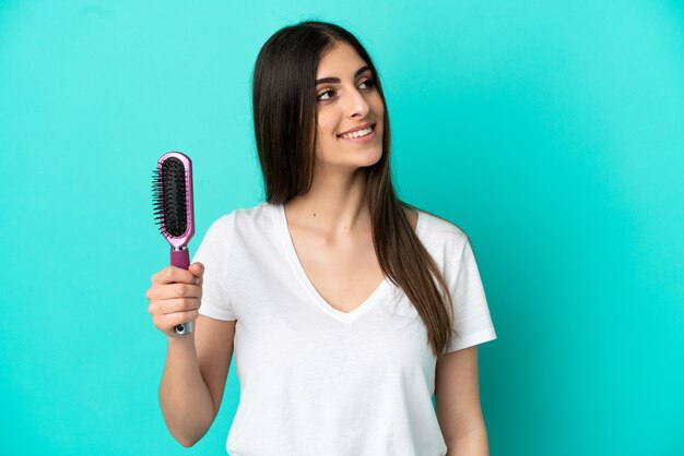 Young caucasian woman with hair comb isolated on blue background looking up while smiling