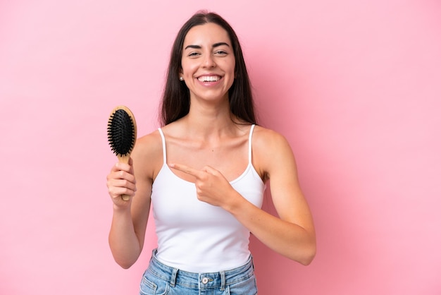 Young caucasian woman with hair comb isolated on pink\
background and pointing it