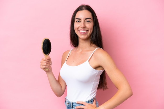 Young caucasian woman with hair comb isolated on pink background smiling a lot