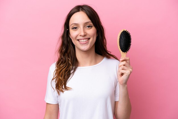 Young caucasian woman with hair comb isolated on pink background smiling a lot