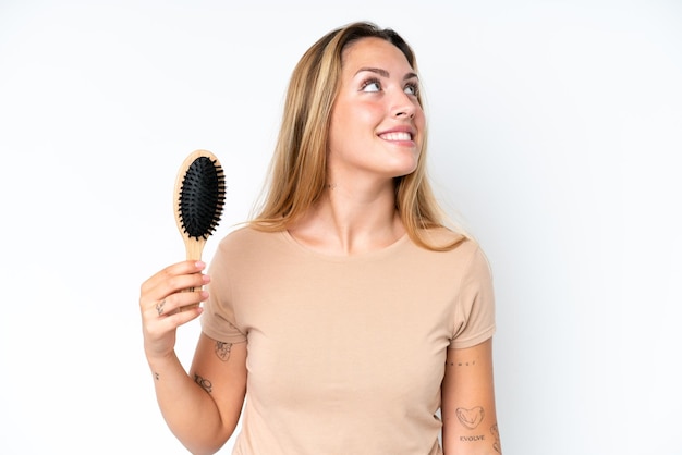 Young caucasian woman with hair comb isolated on white background looking up while smiling