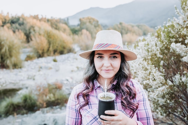 Young caucasian woman with a hat and wearing a pale pink blouse drinking mate in a natural space