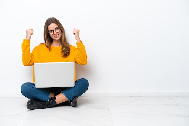 Young caucasian woman with a laptop sitting on the floor isolated on white background celebrating a victory