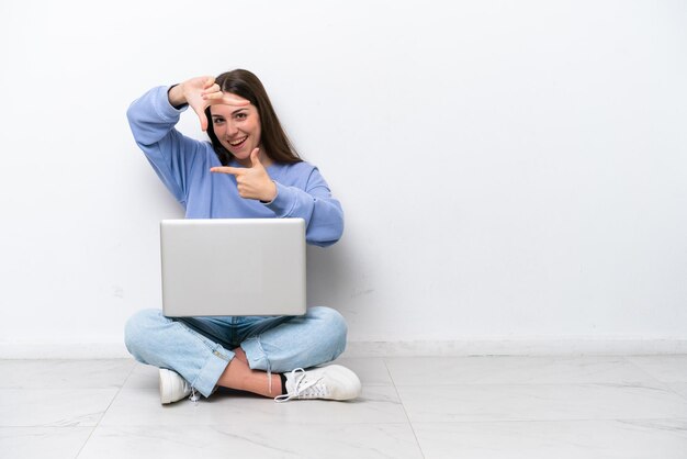 Young caucasian woman with laptop sitting on the floor isolated on white background focusing face Framing symbol