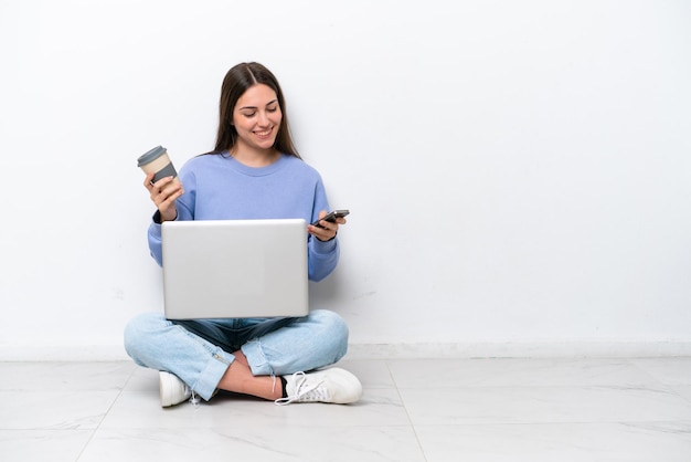 Young caucasian woman with laptop sitting on the floor isolated on white background holding coffee to take away and a mobile