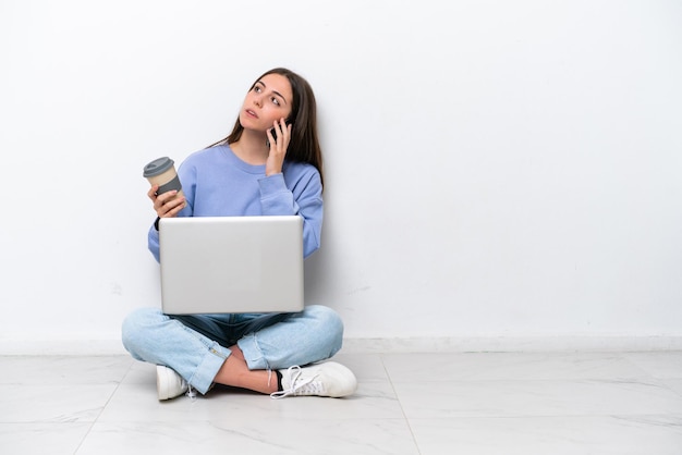 Young caucasian woman with laptop sitting on the floor isolated on white background holding coffee to take away and a mobile