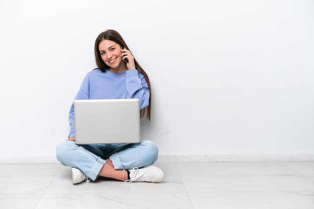 Young caucasian woman with laptop sitting on the floor isolated on white background keeping a conversation with the mobile phone with someone