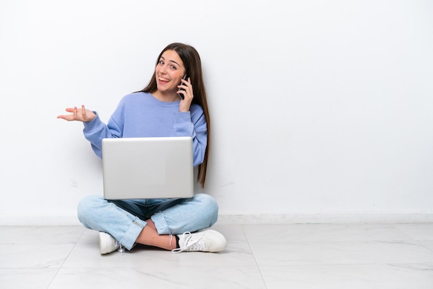 Young caucasian woman with laptop sitting on the floor isolated on white background keeping a conversation with the mobile phone with someone
