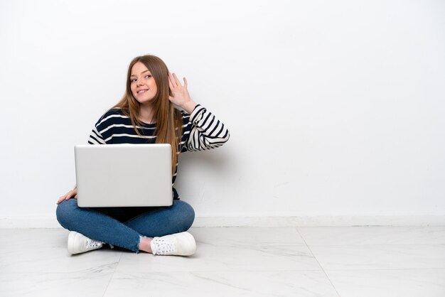 Young caucasian woman with a laptop sitting on the floor isolated on white background listening to something by putting hand on the ear