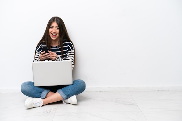 Young caucasian woman with a laptop sitting on the floor isolated on white background surprised and sending a message