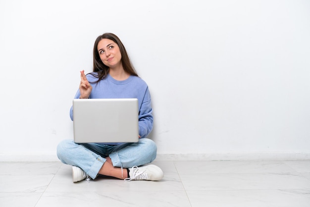 Giovane donna caucasica con laptop seduto sul pavimento isolato su sfondo bianco con le dita incrociate e augurando il meglio