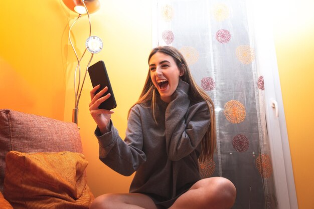 リビングルームでスマートフォンを使用しながら笑っている若い白人女性。