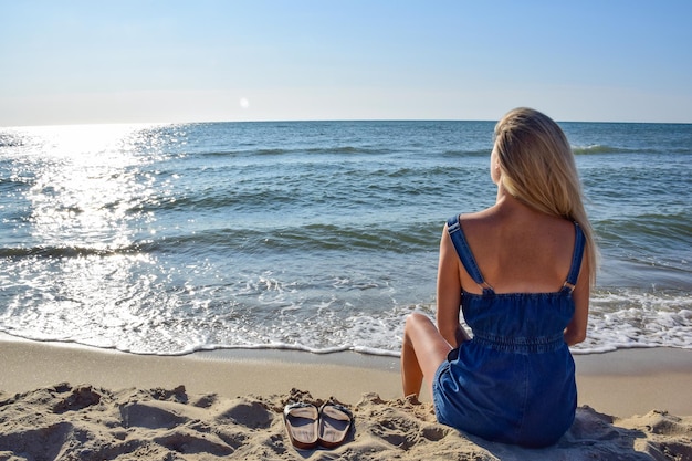 長い白い髪の若い白人女性がビーチに座って海を見ている背面図