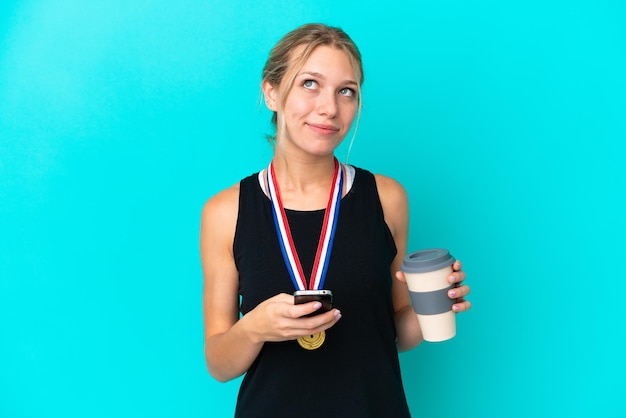 奪うためにコーヒーと何かを考えている間携帯電話を保持している青い背景に分離されたメダルを持つ若い白人女性