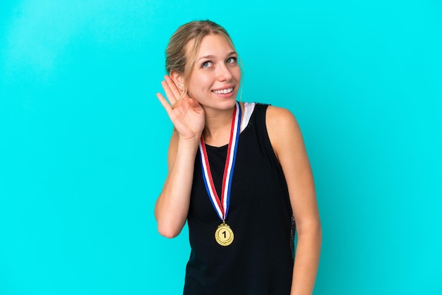 Young caucasian woman with medals isolated on blue background listening to something by putting hand on the ear