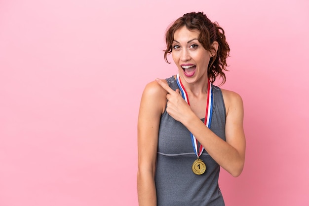 Young caucasian woman with medals isolated on pink background surprised and pointing side