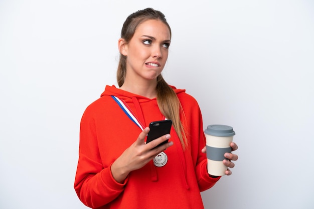 Young caucasian woman with medals isolated on white background holding coffee to take away and a mobile while thinking something