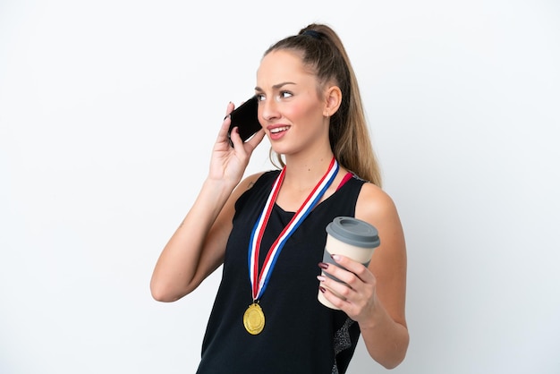 Молодая кавказская женщина с медалями на белом фоне держит кофе на вынос и мобильный телефон