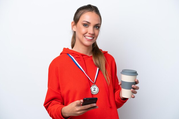 Young caucasian woman with medals isolated on white background holding coffee to take away and a mobile