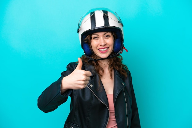 Young caucasian woman with a motorcycle helmet isolated on blue\
background with thumbs up because something good has happened