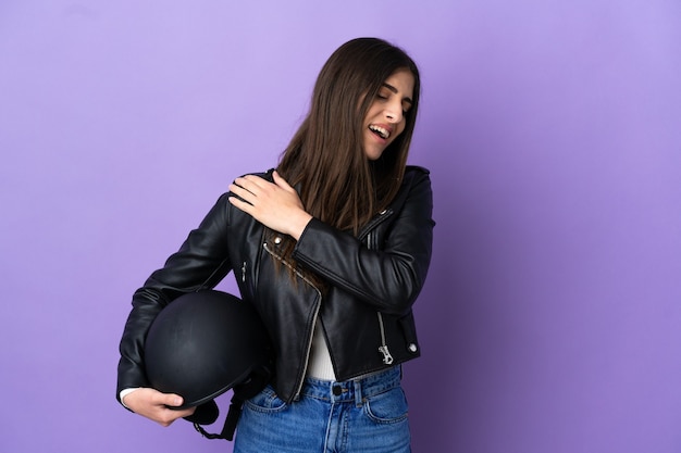 Молодая кавказская женщина в мотоциклетном шлеме изолирована на фиолетовом фоне и страдает от боли в плече за то, что приложила усилие