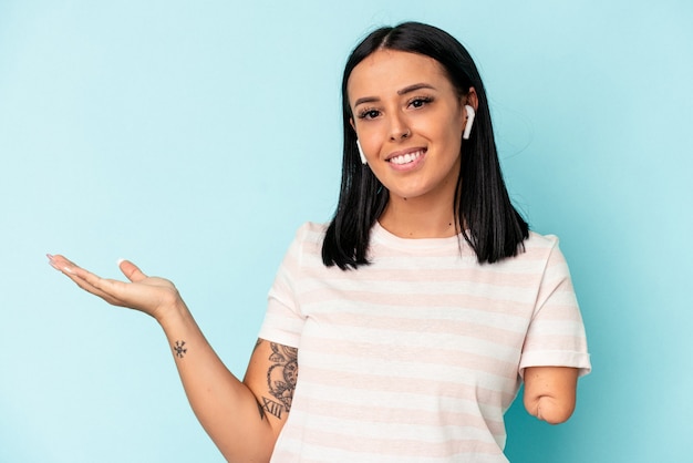 Young caucasian woman with one arm listening to music with wireless headphones isolated on blue background showing a copy space on a palm and holding another hand on waist.