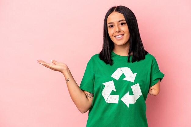 Young caucasian woman with one arm recycled waste isolated on pink background showing a copy space on a palm and holding another hand on waist.
