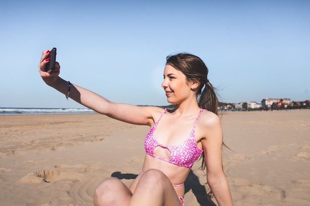 Молодая кавказская женщина с розовым бикини на пляже с помощью мобильного телефона